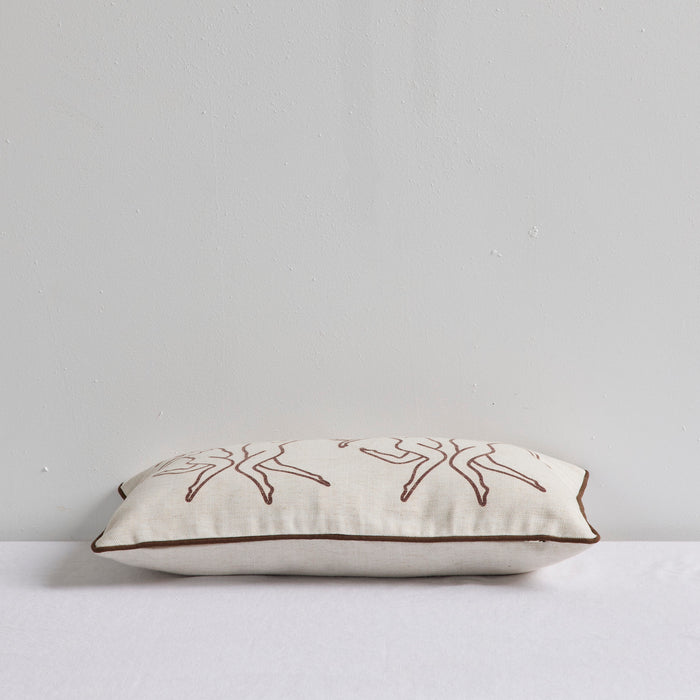 A rectangular cream linen throw pillow with brown embroidered line-art figures and a brown piped edge, resting on a white surface against a light gray wall.