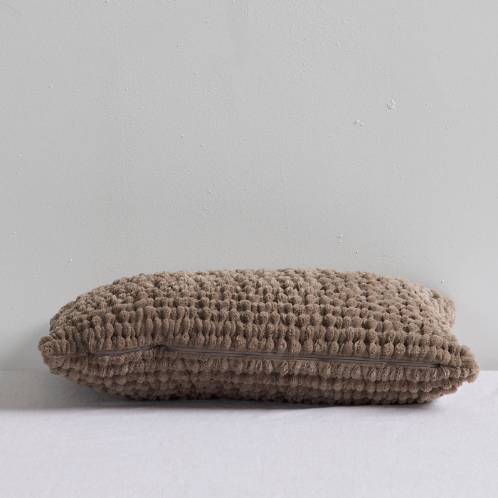A textured, chunky knit throw pillow in a taupe-gray color, resting on a white surface against a light gray wall.
