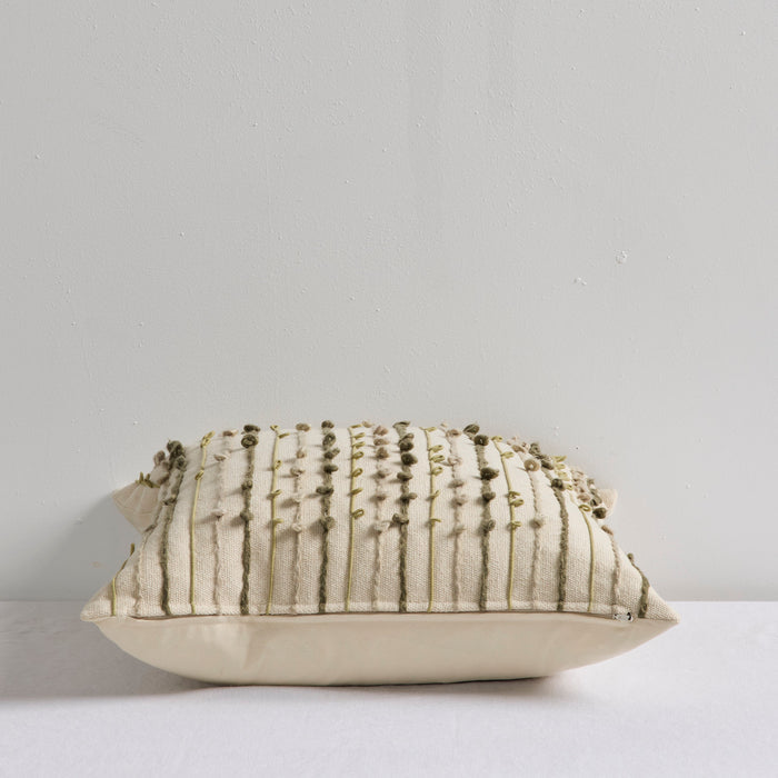 A square decorative throw pillow in cream fabric with vertical olive green textured stripes and small hanging bead accents, resting on a white surface against a light gray wall.