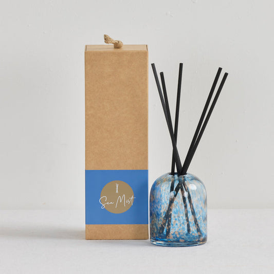 Aurora Diffuser - Sea Mist reed diffuser in a mottled blue and gold glass vessel beside kraft gift box