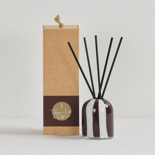 Aurora Diffuser - Tobacco Leaf & Saffron reed diffuser in an organic striped glass vessel with black reeds beside a kraft gift box