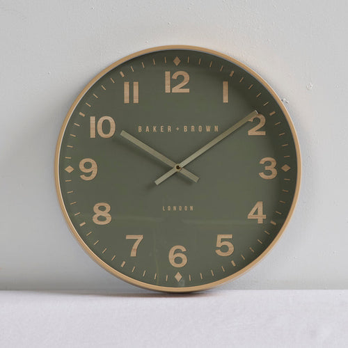 A round wall clock with a muted olive green face, gold-toned numerals and frame, and silver hands, shown against a light gray wall.