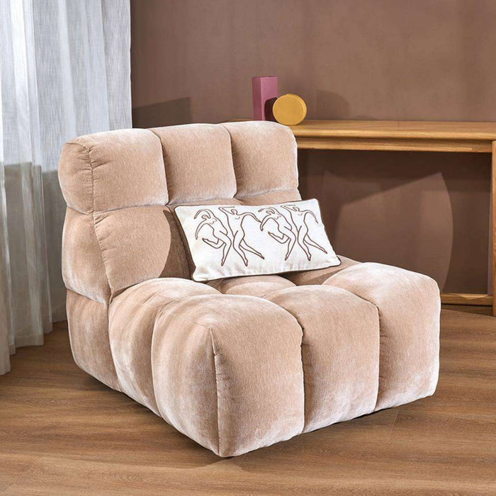 A beige plush modular foam chair with a white accent pillow featuring a black line-art illustration of three figures, set in a living room environment with a wooden floor, brown wall, and a console table in the background.