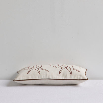 A rectangular cream linen throw pillow with brown embroidered line-art figures and a brown piped edge, resting on a white surface against a light gray wall.