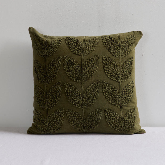 An olive green throw pillow with a textured, raised leaf pattern, set against a plain light gray wall and white surface.