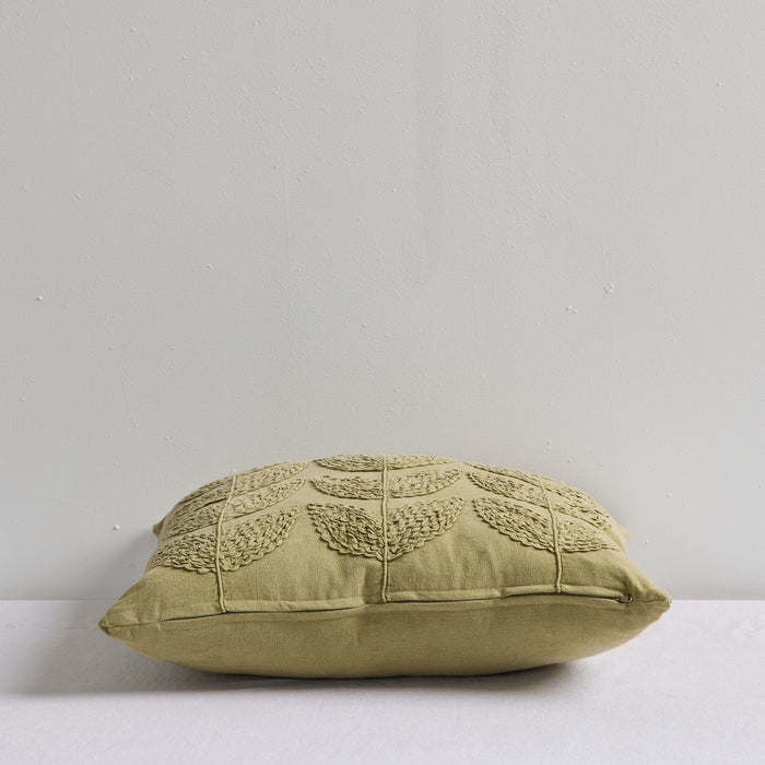 An olive green decorative throw pillow with a textured, raised leaf pattern on the front, resting on a white surface against a light gray wall.