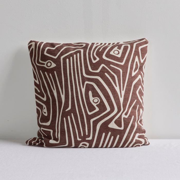 A square throw pillow with an abstract brown and cream line pattern, resting on a white surface against a light gray wall.
