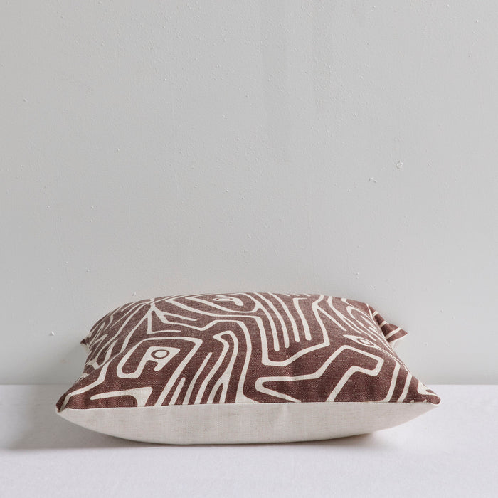 A square throw pillow with an abstract brown and cream topographic line pattern, resting on a white surface against a light gray wall.