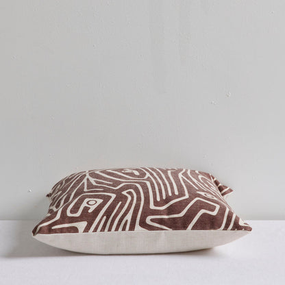 A square throw pillow with an abstract brown and cream topographic line pattern, resting on a white surface against a light gray wall.