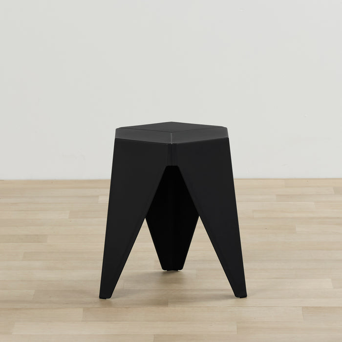 A modern black geometric stool or side table with a triangular three-legged base, shown on a light wood floor against a plain white wall.