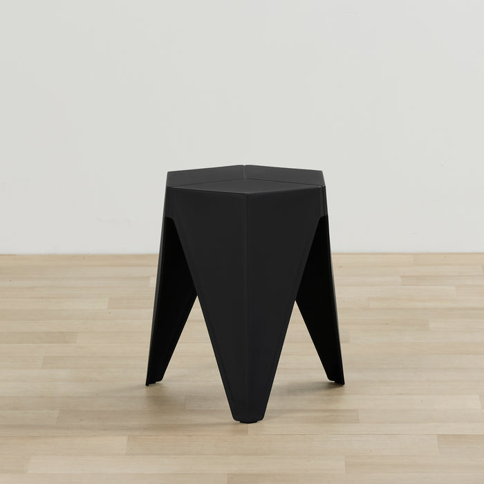 A modern black geometric stool or side table with a triangular three-legged base, shown on a light wood floor against a plain white wall.