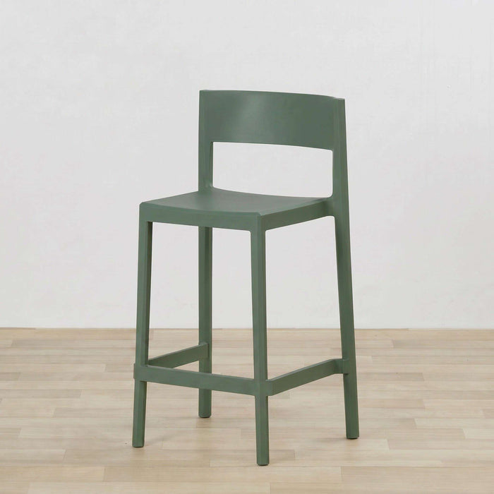 A moderngreen bar stool with a minimalist design, featuring a curved backrest, a flat seat, and a lower support rail, set against a plain white wall and light wood floor.