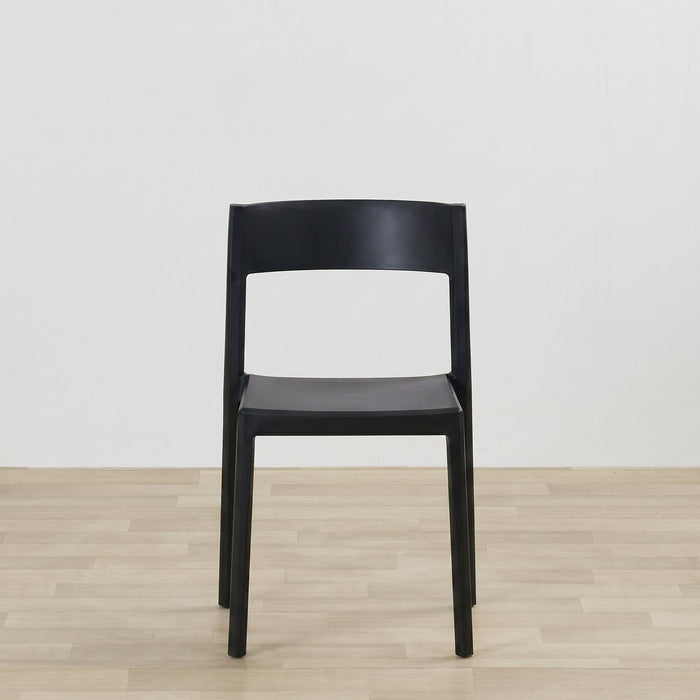 A minimalist black molded plastic dining chair shown from the front, set against a plain white wall and light wood floor.