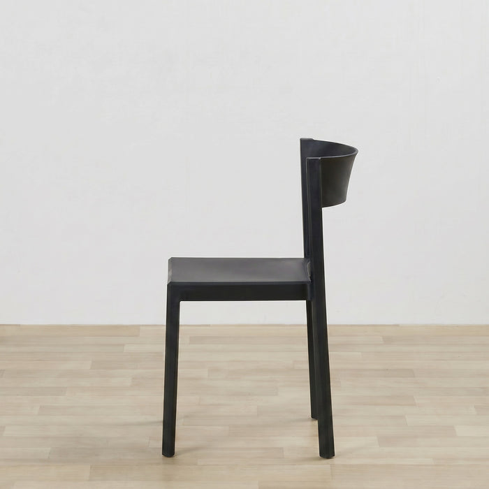 A minimalist black dining chair with a sculptural, integrated backrest, shown from a side profile against a white wall and light wood floor.
