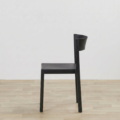 A minimalist black dining chair with a sculptural, integrated backrest, shown from a side profile against a white wall and light wood floor.