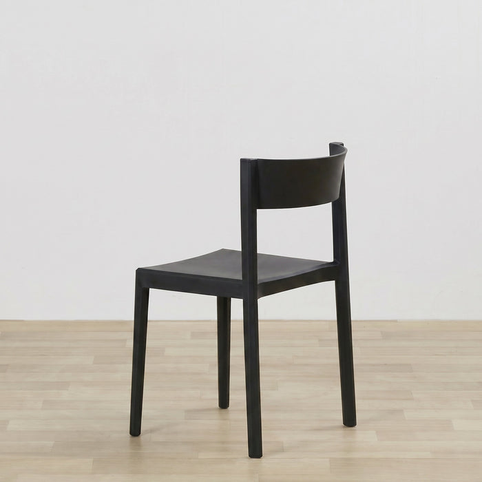 A minimalist black wooden dining chair with a curved backrest and square seat, shown from a three-quarter angle on a light wood floor against a white wall.