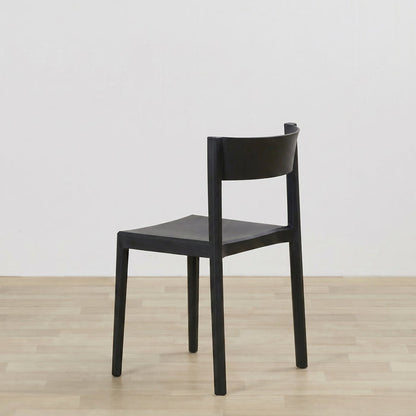 A minimalist black wooden dining chair with a curved backrest and square seat, shown from a three-quarter angle on a light wood floor against a white wall.
