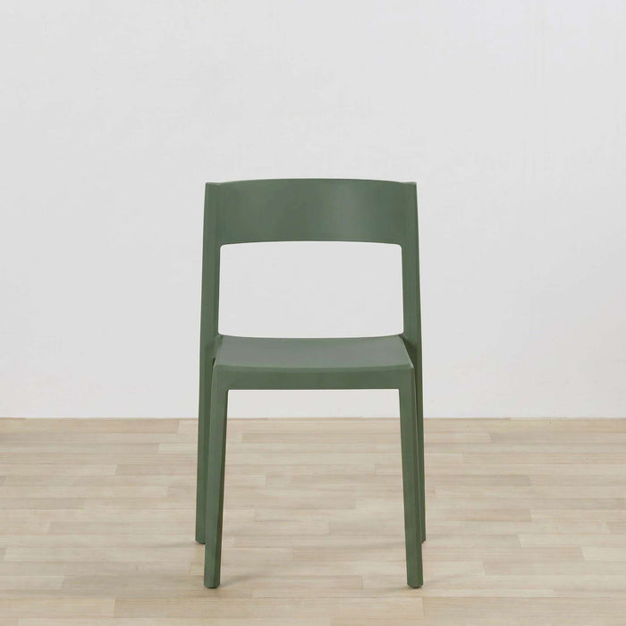 A modern molded plastic chair in a muted sage green color, positioned centrally against a white wall and light wood floor.