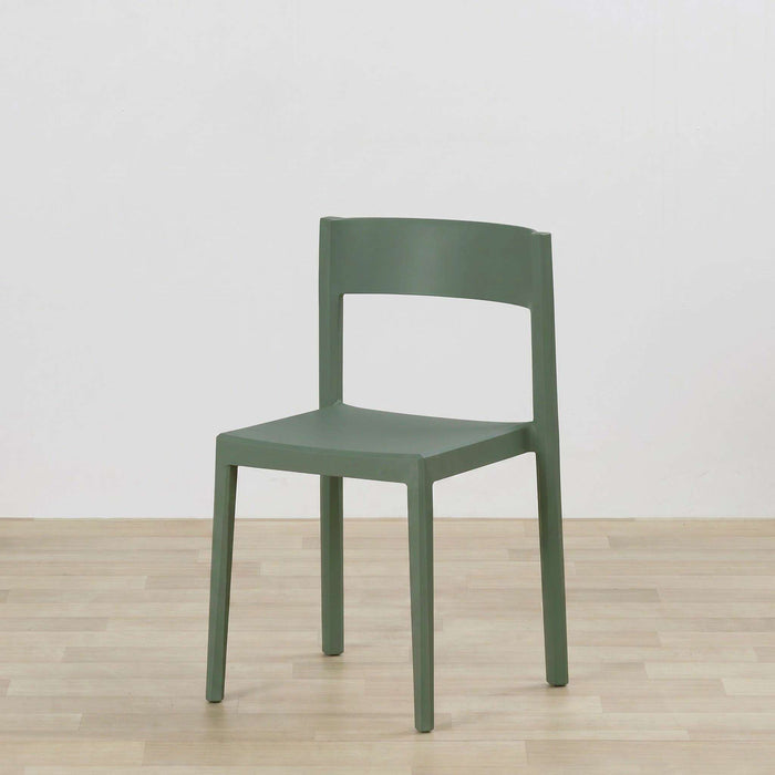 A modern side chair in a muted sage green color, positioned on a light wood floor against a plain white wall.