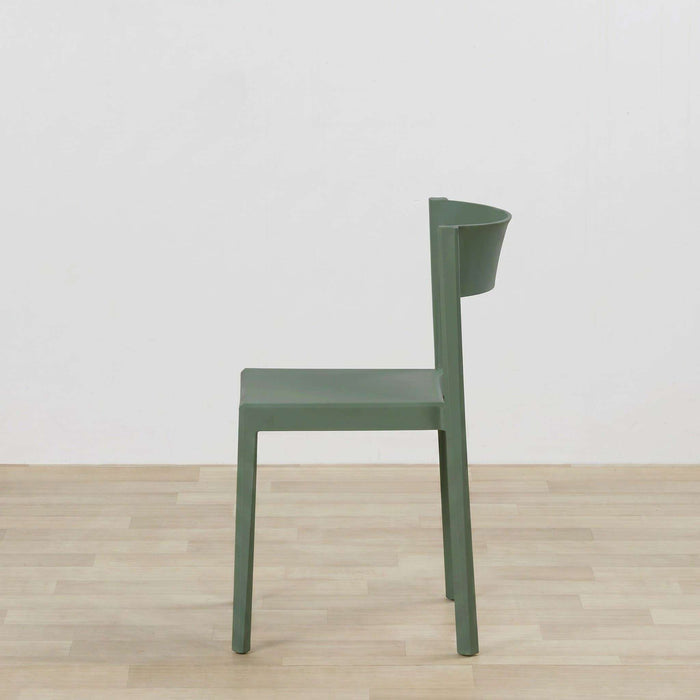 A modern molded plastic dining chair in a muted sage green color, positioned on a light wood floor against a plain white wall.