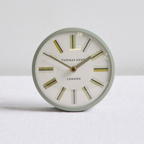 A round Thomas Kent London wall clock with a light sage green frame, white dial, and gold-toned hour markers and hands, set against a neutral light gray background.