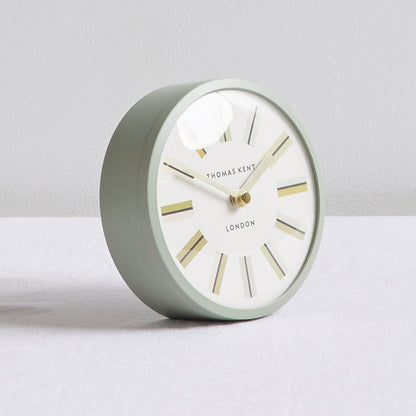 A round Thomas Kent London table clock with a matte sage green body, white dial, and gold-toned hands and hour markers, set against a neutral light gray background.