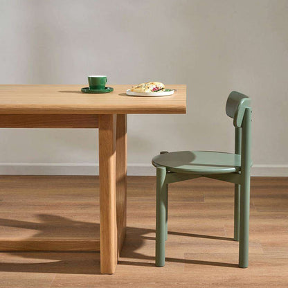 A light natural wood dining table with a matching green chair in a minimalist room setting, featuring a green cup and a plate of food on the tabletop.