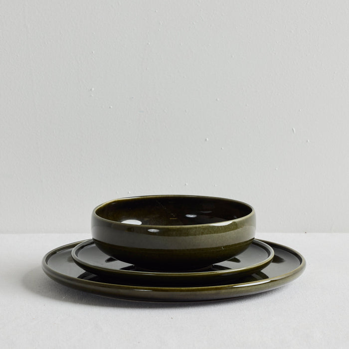 A dark olive green ceramic dinnerware set consisting of a bowl and two matching plates, stacked neatly on a light gray surface against a neutral gray background.