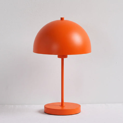 A minimalist orange table lamp with a dome-shaped shade, a single central stem, and a round base, photographed against a plain light gray wall and white surface.