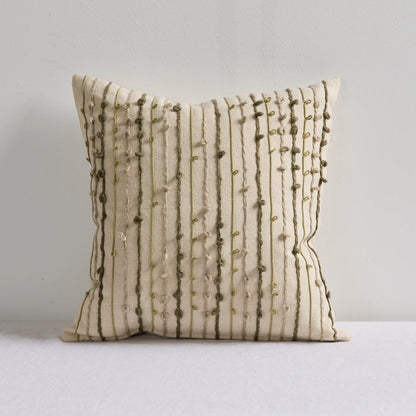A square throw pillow in cream fabric with vertical olive green embroidered stripes and bead accents, resting on a white surface against a light gray wall.