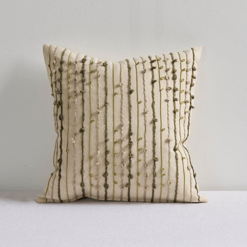 A square throw pillow in cream fabric with vertical olive green embroidered stripes and bead accents, resting on a white surface against a light gray wall.