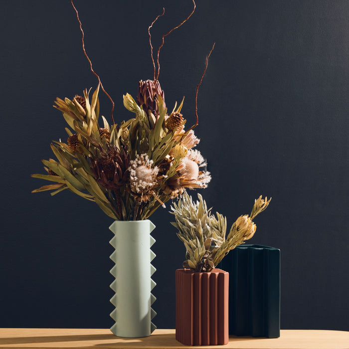 Stria Square Vase - Brick, brick-coloured ribbed vase with dried foliage, styled beside tall sculptural vases on a timber surface