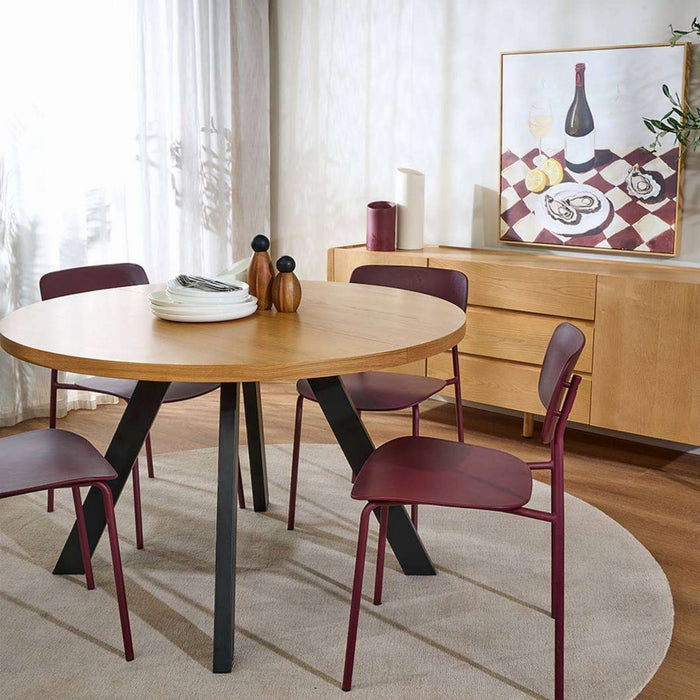 A round wooden dining table with black angled legs, surrounded by four modern chairs with burgundy seats and backs, set on a light beige rug in a bright dining room.